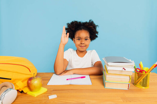 Happy African American schoolgirl sitting at desk in class and stretching hand uphill on blue background. Back to school concept.