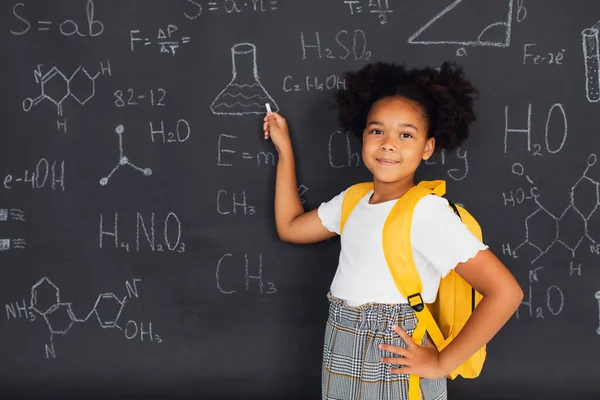 Happy African American schoolgirl solving problems near the blackboard at school, back to school concept.