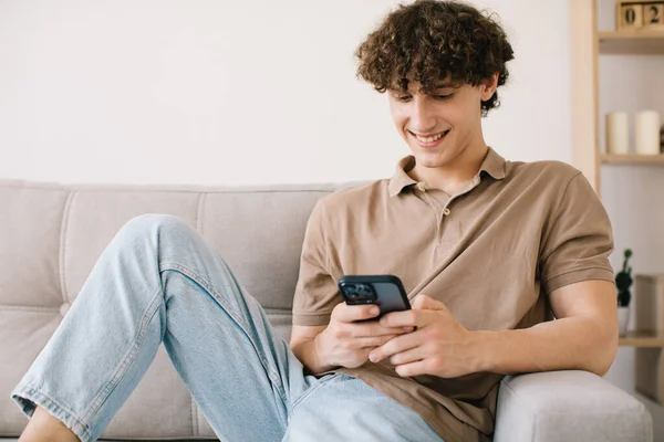年轻快乐的卷发男子坐在客厅沙发上休息时使用智能手机的画像 — 图库照片