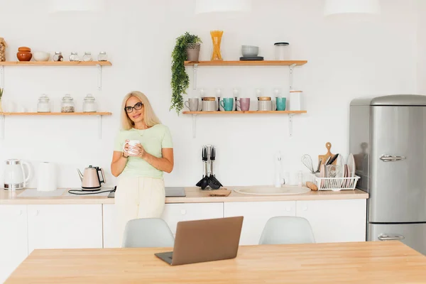 Modern attractive middle-aged woman drinking coffee and working on laptop in kitchen at home