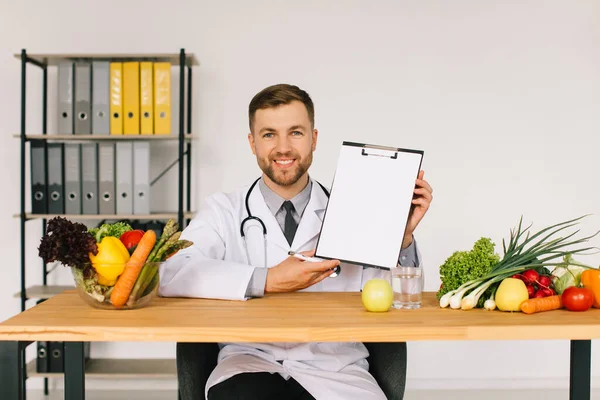 Happy doctor nutritionist sitting at workplace at desk in office among fresh vegetables and showing folder with copy space, diet plan concept