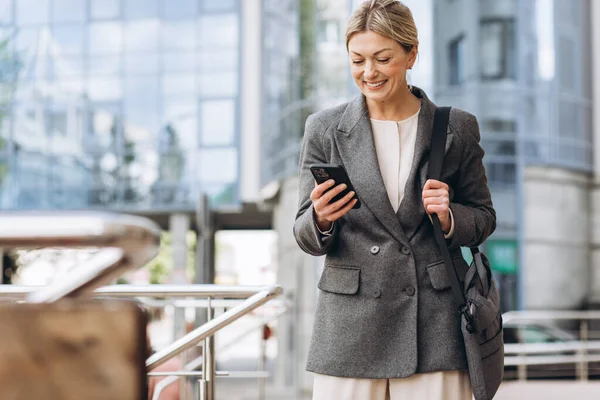 一个穿着西服和灰色夹克的漂亮而成熟的女商人的画像 在现代都市背景下微笑着和电话交谈 — 图库照片