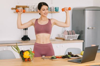 Kollarını dambıllarla gösteren mutlu spor kadın blogcusu portresi ve mutfakta dizüstü bilgisayarda sağlıklı beslenme üzerine video konferansı.