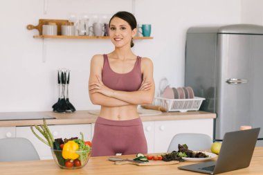 Mutlu spor kadın blog yazarı portresi taze sebzelerle masada, sağlıklı beslenme konseptiyle.
