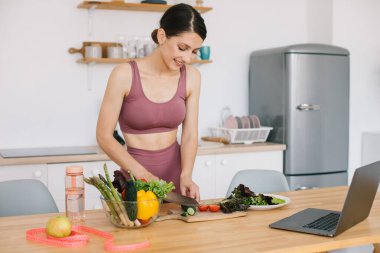 Mutlu atletik kadın blogcu beslenme uzmanı taze sebzeli bir salata hazırlıyor ve mutfaktaki dizüstü bilgisayarda sağlıklı beslenmeyle ilgili bir video konferansı düzenliyor.