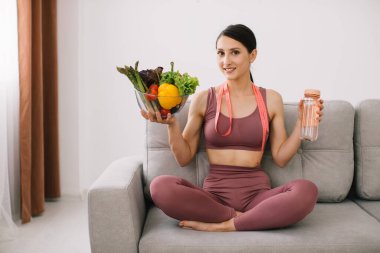 Kanepede oturan genç sporcu kadın elinde çeşitli sebzeler, bir şişe su ve paket cetvel, kilo kaybı ve sağlıklı beslenme kavramları tutuyordu..