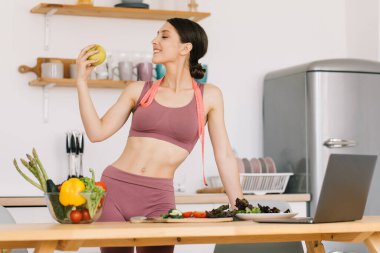 Mutlu spor kadının portresi taze elma tutuyor ve bisepsleri mutfakta gösteriyor, sağlıklı beslenme konsepti.