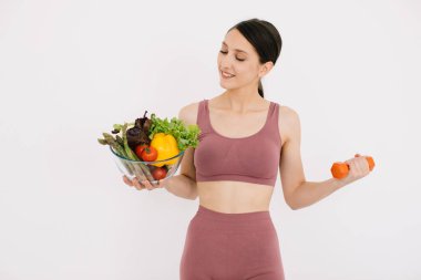 Çeşitli sağlıklı sebzelerle dolu tepsisiyle güzel mutlu genç bir kadın ve beyaz arka planda izole edilmiş halterli kaslarını gösteriyor.