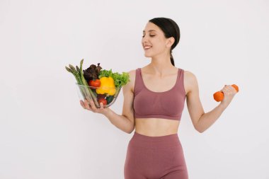 Çeşitli sağlıklı sebzelerle dolu tepsisiyle güzel mutlu genç bir kadın ve beyaz arka planda izole edilmiş halterli kaslarını gösteriyor.