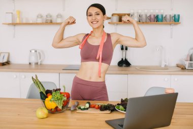 Bisepsleri gösteren mutlu spor kadın blogcusu portresi ve mutfaktaki dizüstü bilgisayarda sağlıklı beslenme üzerine video konferansı.