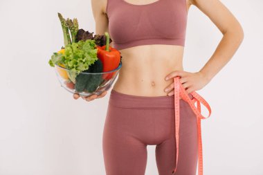 Elinde farklı sebzeler ve beyaz arka planda cetvel olan mutlu bir kadın.