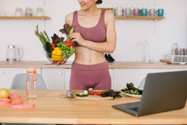 Athletic woman blogger nutritionist prepare a salad with fresh vegetables and conducts a video conference on healthy eating on laptop in the kitchen