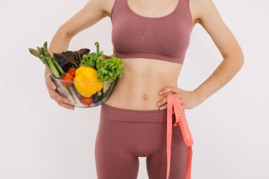 Elinde farklı sebzeler ve beyaz arka planda cetvel olan mutlu bir kadın.