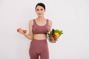 Çeşitli sağlıklı sebzelerle dolu tepsisiyle güzel mutlu genç bir kadın ve beyaz arka planda izole edilmiş halterli kaslarını gösteriyor.