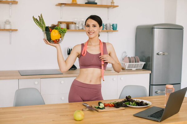 Портрет счастливой спортивной женщины-блогера с тарелкой свежих овощей и ведущей видеоконференцией по здоровому питанию на ноутбуке на кухне