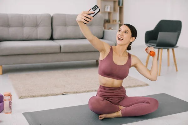 stock image Attractive young woman taking selfie showing biceps with dumbbell at home.