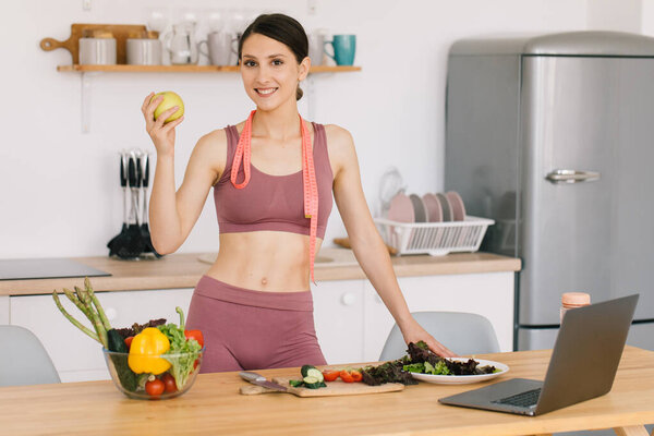 Portrait of happy sporty woman holding fresh apple and showing biceps in kitchen, healthy eating concept