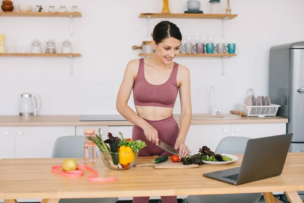 stock image Happy athletic woman blogger nutritionist prepare a salad with fresh vegetables and conducts a video conference on healthy eating on laptop in the kitchen