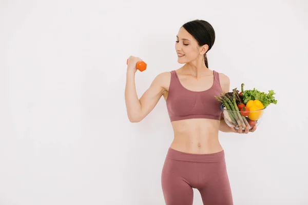 様々な健康的な野菜のトレイを持つ美しい幸せな若い女性と白い背景に隔離されたダンベルで彼女の筋肉を示す — ストック写真