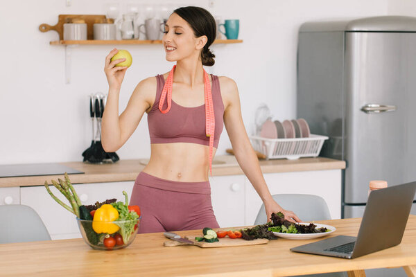Портрет счастливой спортивной женщины, держащей свежее яблоко и показывающей бицепсы на кухне, концепция здорового питания