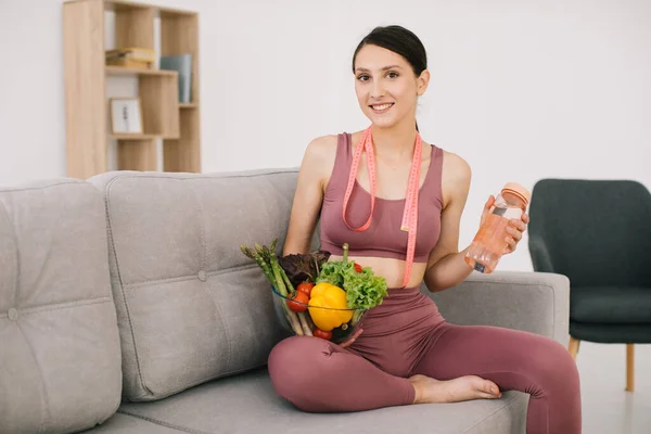 年轻的女运动员坐在沙发上 手里拿着各种各样的蔬菜 瓶装水和包裹着的尺子 体重减轻和健康饮食的理念 — 图库照片