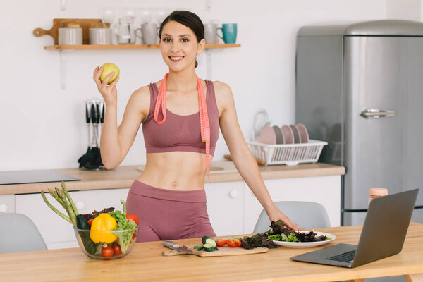 Портрет счастливой спортивной женщины, держащей свежее яблоко и показывающей бицепсы на кухне, концепция здорового питания