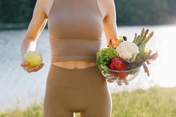 Female vegan holding apple and plate of fresh vegetables near the belly in outdoors