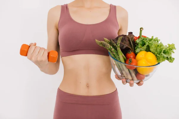 美丽而快乐的年轻女子 她端着一盘盘健康的蔬菜 用白色背景上的哑铃展示她的肌肉 — 图库照片