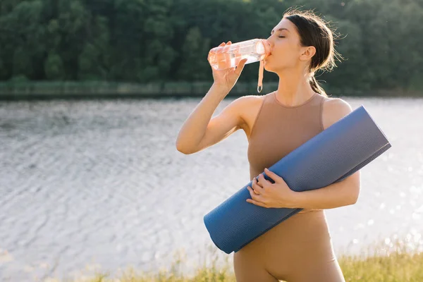 Athletic woman drinks fresh water and holds a yoga mat on the outdoors