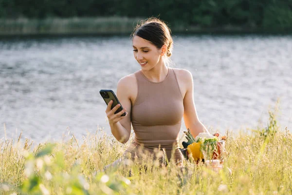 Mujer Deportiva Feliz Mirando Teléfono Sosteniendo Verduras Frescas Aire Libre — Foto de Stock