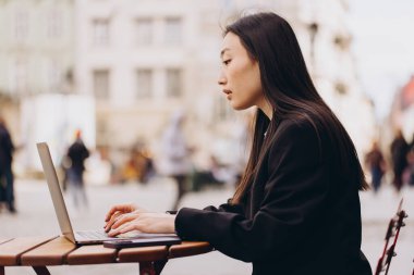 İş için serbest çalışan genç Asyalı bir kadın dizüstü bilgisayarını iş için kullanıyor ve telefonla konuşuyor. Gündüz açık havada oturmak. Modern teknolojiye sahip şehir halkı yaşam tarzı.