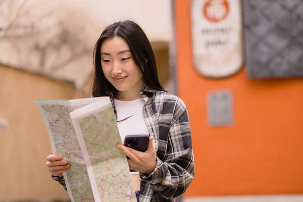 一个漂亮的黑发韩国女人拿着地图的肖像 并将其与旧城街道上的智能手机导航相比较 在欧洲旅行的亚洲女游客 — 图库照片