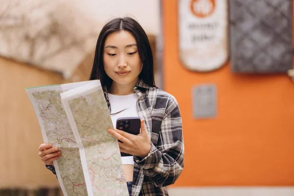一个漂亮的黑发韩国女人拿着地图的肖像 并将其与旧城街道上的智能手机导航相比较 在欧洲旅行的亚洲女游客 — 图库照片