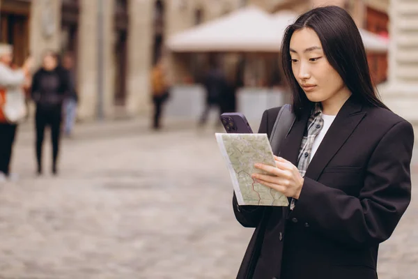 地図やスマートフォンのナビゲーションで方向を確認しながら ヨーロッパの古い街の通りにカジュアルな服で若い幸せなアジアの女性の観光客を肖像画 — ストック写真