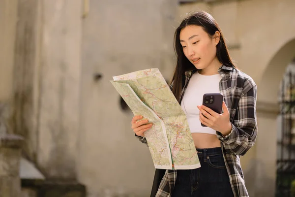 美しいブルネットの韓国人女性の肖像画地図を保持し 旧市街の通りにスマートフォンのナビゲーションと比較してください ヨーロッパを旅するアジアの観光客の女性 — ストック写真