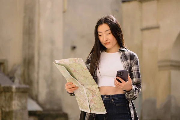 美しいブルネットの韓国人女性の肖像画地図を保持し 旧市街の通りにスマートフォンのナビゲーションと比較してください ヨーロッパを旅するアジアの観光客の女性 — ストック写真