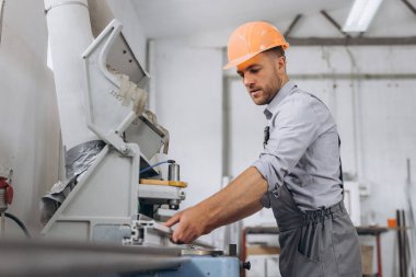 Özel üniformalı ve turuncu kasklı bir erkek işçi fabrikadaki PVC pencere ve kapı üretimi için CNC makinesinde çalışıyor..