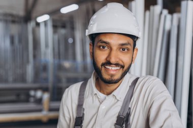 Beyaz kasketli ve tulumlu mutlu bir Hindu işçinin portresi bir fabrika ve alüminyum çerçevelerin arka planına karşı hidrolik bir kamyon tutuyor..