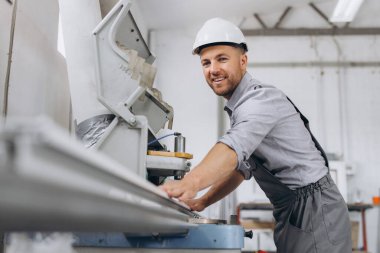 Özel üniformalı ve beyaz kasklı bir erkek işçi fabrikadaki PVC pencere ve kapı üretimi için CNC makinesinde çalışıyor..