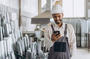 Özel üniformalı mutlu Hintli erkek fabrika işçisi ve telefonu tutan beyaz başlıklı adam metal-plastik pencere ve kapı üretiminde diyalog kuruyor.