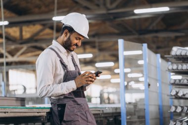 Özel üniformalı mutlu Hintli erkek fabrika işçisi ve telefonu tutan beyaz başlıklı adam metal-plastik pencere ve kapı üretiminde diyalog kuruyor.