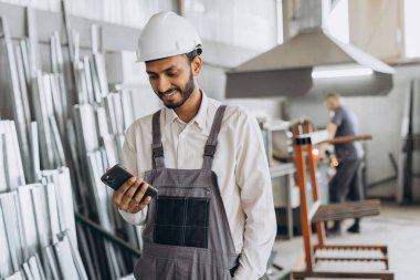 Özel üniformalı mutlu Hintli erkek fabrika işçisi ve telefonu tutan beyaz başlıklı adam metal-plastik pencere ve kapı üretiminde diyalog kuruyor.