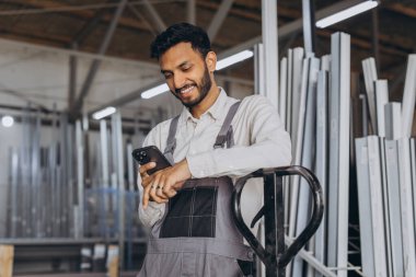 Portrait of a happy Hindu worker in a white helmet and overalls holding a hydraulic truck and talking on the phone against a background of a factory and aluminum frames. clipart