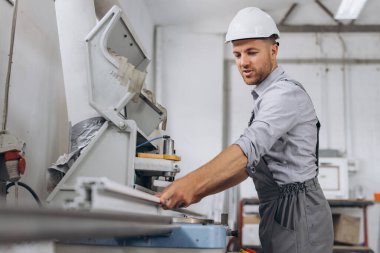 Özel üniformalı ve beyaz kasklı bir erkek işçi fabrikadaki PVC pencere ve kapı üretimi için CNC makinesinde çalışıyor..