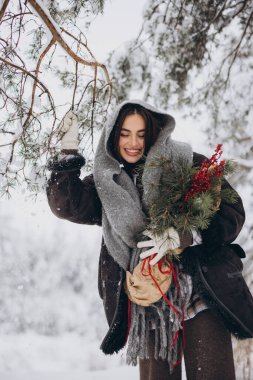 Örgü atkı ve eldiven takmış, elinde çam buketi tutan ve karlı ormanda poz veren mutlu güzel bir kadının portresi.