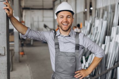 Gri üniformalı mutlu bir işçinin portresi ve fabrika yapımının arka planında poz veren beyaz bir şapka.