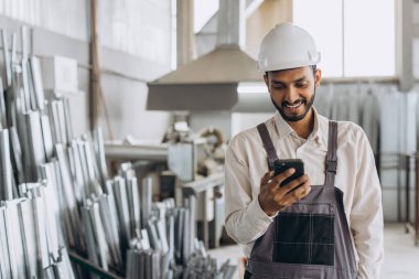 Özel üniformalı mutlu Hintli erkek fabrika işçisi ve telefonu tutan beyaz başlıklı adam metal-plastik pencere ve kapı üretiminde diyalog kuruyor.