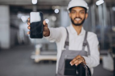 Beyaz şapkalı ve tulumlu mutlu Hindu işçilerin portresi akıllı telefon tutuyor ve fabrika arka planında boş ekran gösteriyor..