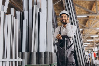 Portrait of a happy Hindu worker in a white hard hat and overalls holding a hydraulic truck against a background of a factory and aluminum frames. clipart