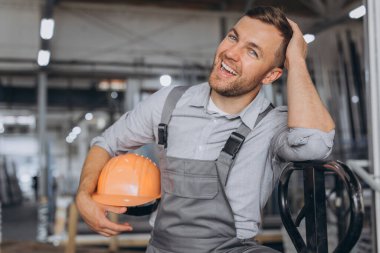 Turuncu şapkalı ve tulumlu mutlu bir işçinin portresi bir fabrika ve alüminyum çerçevelerin arka planında hidrolik bir kamyon tutuyor..
